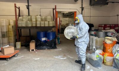 An investigator in a hazmat suit stands with paraphernalia suspected of use in making illicit drugs