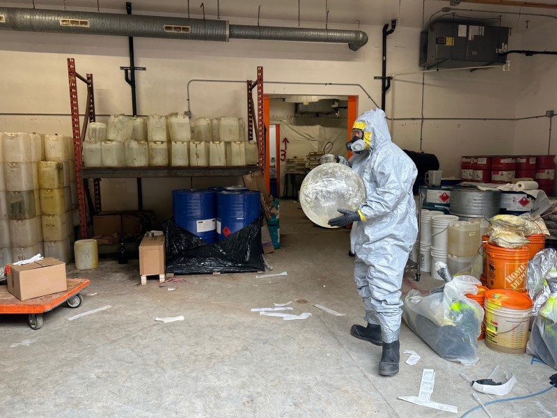 An investigator in a hazmat suit stands with paraphernalia suspected of use in making illicit drugs