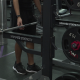 A person is seen at a gym lining up to use a squat rack, with a weight filled bar resting on the rack.