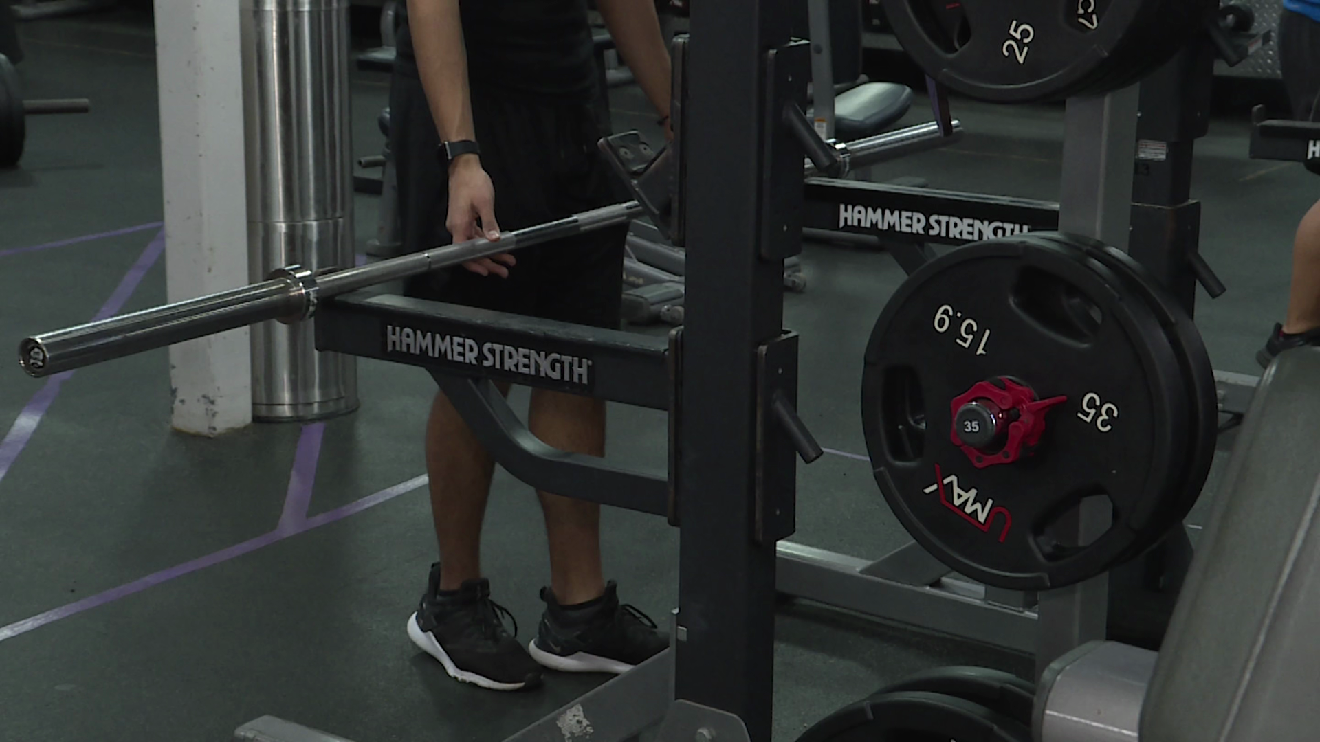 A person is seen at a gym lining up to use a squat rack, with a weight filled bar resting on the rack.